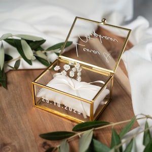Könnte beinhalten: Eine transparente Glas-Ringbox mit goldenem Metallrahmen, geöffnet, um ein weißes Satin-Kissen und kleine weiße Blumen zu zeigen. Die Box trägt handschriftlichen Text und das Datum 25.10.2023. Olivenzweige sind im Vordergrund.