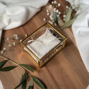 Op de afbeelding: Een rechthoekige glazen ringdoos met een gouden frame en een wit kussen. De namen "Lena" en "Stephan" en de datum "28.10.2022" staan op het glas. De doos ligt op een houten oppervlak, met witte stof en groen in de achtergrond.