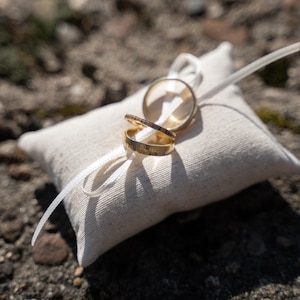 May include: Two gold wedding bands on a white pillow with a white ribbon.