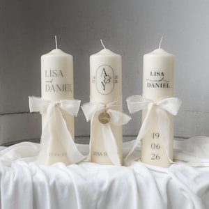 Könnte beinhalten: Drei elfenbeinfarbene Säulenkerzen mit schwarzem Text und cremefarbenen Bändern. Die Kerzen sind mit Namen, Daten und einem Monogramm personalisiert. Eine Kerze trägt die Aufschrift "LISA and DANIEL" und "21/06/20". Eine andere trägt "A & Y" und "ANNA & 20 26". Die letzte trägt "LISA DANIEL" und "19 06 26".