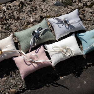 May include: Six small, rectangular, fabric ring bearer pillows in various colors, including white, light blue, pink, and green. Each pillow has a satin ribbon and a pair of rings on top.