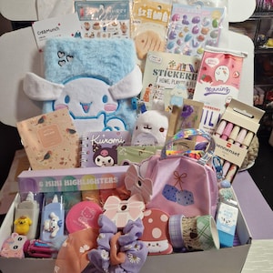 Könnte beinhalten: Eine offene Box voller Schreibwaren und Accessoires. Sie enthält ein blaues Plüsch-Notizbuch mit einer Cartoonfigur, Notizbücher, Aufkleber, Stifte, Textmarker, eine rosa Tasche und dekorative Klebebänder. Die Box ist mit farbenfrohen Artikeln gefüllt.