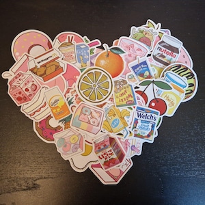 Puede incluir: Una colección de pegatinas coloridas dispuestas en forma de corazón. Las pegatinas presentan varios alimentos como rosquillas, naranjas y cajas de jugo. Algunas pegatinas tienen texto, incluyendo "McNuggets", "Nutella" y "Welch's Fruit Snacks".