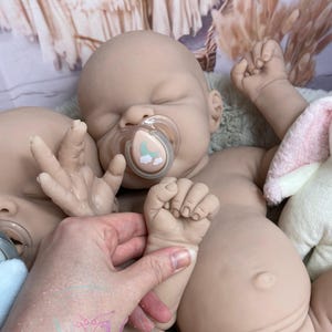 May include: Two realistic baby dolls with closed eyes and pacifiers in their mouths. The dolls are made of soft, lifelike silicone and have detailed features, including tiny fingers and toes.