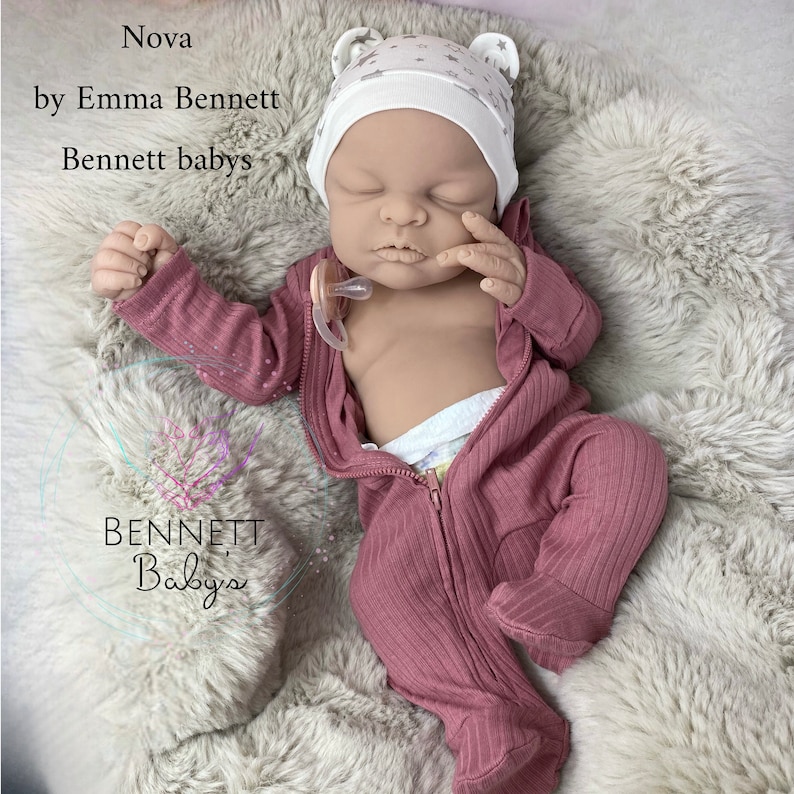 May include: A sleeping reborn baby doll wearing a pink zippered sleeper and a white star patterned hat with ear flaps. The doll is lying on a fluffy white blanket.