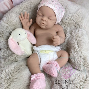 May include: A sleeping reborn baby doll wearing a pink floral hat, a diaper, and pink knitted booties. The doll is lying on a fluffy white blanket with a plush white bunny toy.