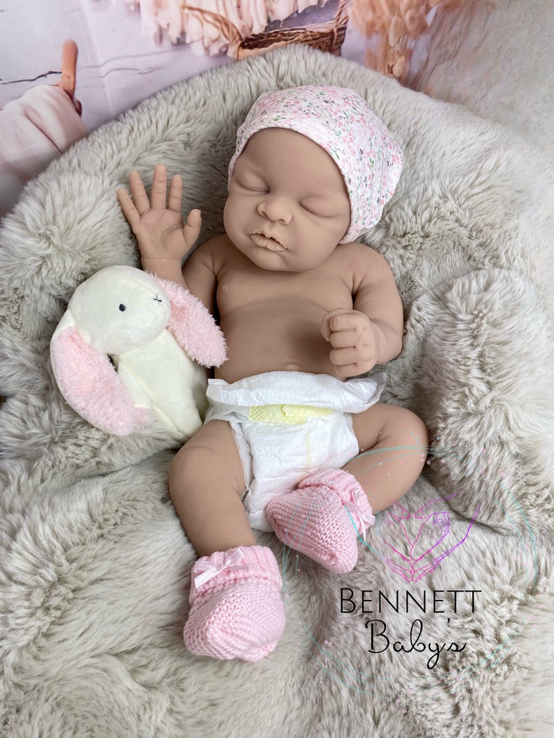 May include: A sleeping reborn baby doll wearing a pink floral hat, a diaper, and pink knitted booties. The doll is lying on a fluffy white blanket with a plush white bunny toy.