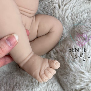 May include: Close-up of a realistic baby doll leg and foot, showing detailed sculpting and realistic skin tone. The foot is slightly curled and the toes are visible.