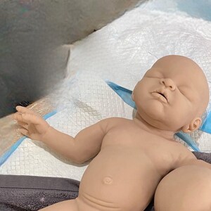 May include: A realistic baby doll with closed eyes and a soft, fleshy appearance. The doll is lying on a white surface with a blue and white patterned fabric.