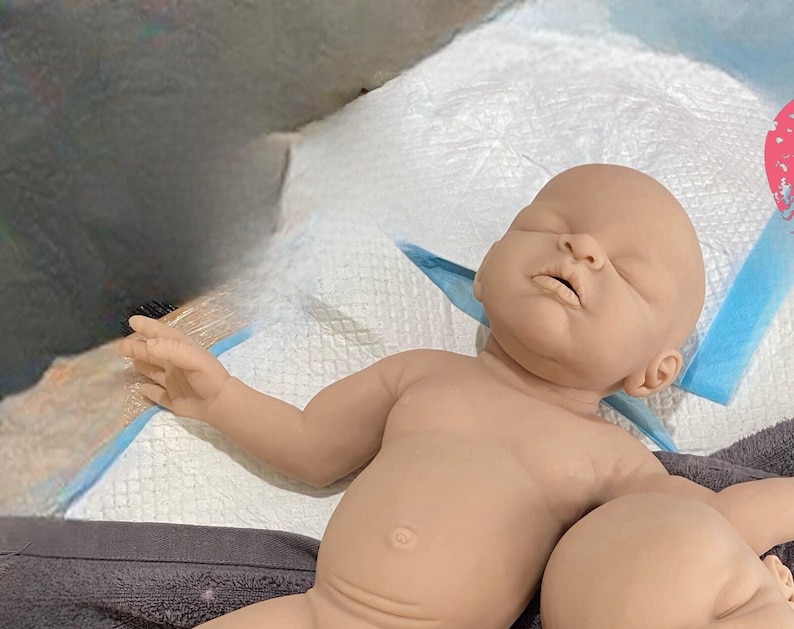 May include: A realistic baby doll with closed eyes and a soft, fleshy appearance. The doll is lying on a white surface with a blue and white patterned fabric.