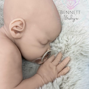 May include: A realistic baby doll with closed eyes, wearing a pacifier, lying on a fluffy white blanket. The doll has a soft, lifelike appearance and is made of silicone.