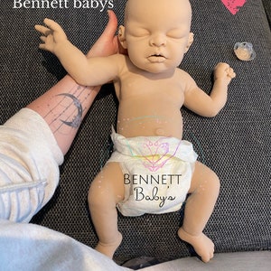 May include: A realistic baby doll with closed eyes, wearing a white diaper with the text "BENNETT Babys" printed on it. The doll is lying on a gray fabric surface.