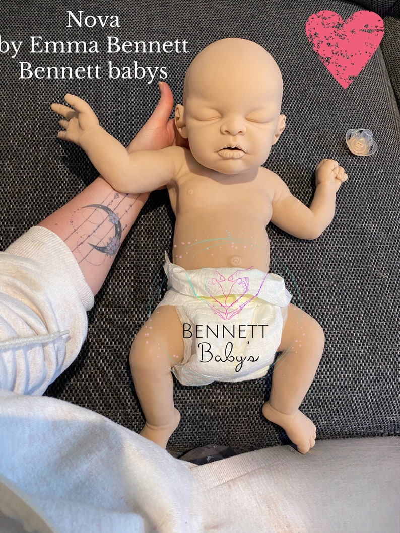 May include: A realistic baby doll with closed eyes, wearing a white diaper with the text "BENNETT Babys" printed on it. The doll is lying on a gray fabric surface.