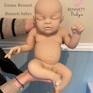 May include: A nude-colored baby doll with closed eyes and a relaxed expression. The doll is being held by a person's hands. The doll is made of soft, realistic-looking silicone. The doll is labeled "Nova by Emma Bennett, Bennett babys".