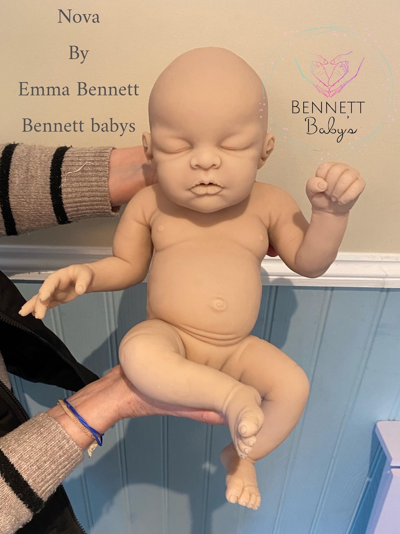 May include: A nude-colored baby doll with closed eyes and a relaxed expression. The doll is being held by a person's hands. The doll is made of soft, realistic-looking silicone. The doll is labeled "Nova by Emma Bennett, Bennett babys".