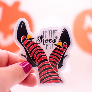 May include: A clear sticker featuring a pair of witch legs in black shoes with red and black striped stockings. The text "IF THE SHOES FIT" is written in black script. The sticker has a Halloween theme.