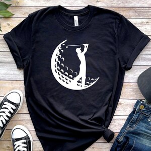 May include: Black t-shirt with a white silhouette of a golfer swinging a club in front of a golf ball.
