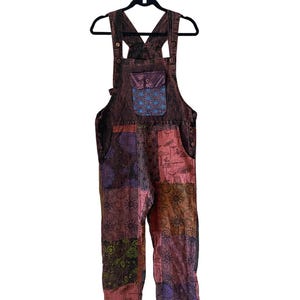 May include: Patchwork denim dungarees with a colourful floral and geometric pattern. The dungarees have a large pocket on the front with a blue and purple design.