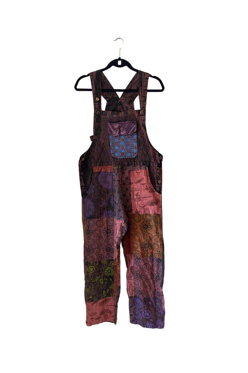 May include: Patchwork denim dungarees with a colourful floral and geometric pattern. The dungarees have a large pocket on the front with a blue and purple design.