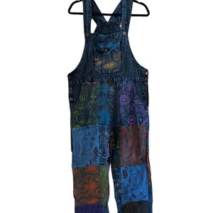 May include: Blue denim dungarees with a patchwork design featuring various colourful patterns. The dungarees have a bib with a pocket and adjustable straps.