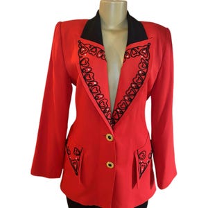 May include: A bright red blazer featuring a black lapel and black embroidered heart-shaped patterns along the lapels and pockets. The jacket includes two gold-toned circular buttons on the front and small clear rhinestones embedded within the black embroidery.