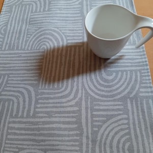 Könnte beinhalten: Eine weiße Keramik-Kaffeetasse mit einem einzigartigen Griff steht auf einem grauen Tischläufer. Der Läufer zeigt ein sich wiederholendes Muster aus weißen, gebogenen Linien und Rechtecken. Der Schatten der Tasse hebt die Textur des Stoffes hervor.