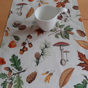 Könnte beinhalten: Eine weiße Keramik-Sahnekännchen steht auf einem cremefarbenen Tischläufer. Der Läufer ist mit Illustrationen von Herbstblättern, Eicheln, Tannenzapfen und Pilzen in Braun-, Orange- und Grüntönen verziert. Der Läufer ist rechteckig.