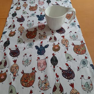 Könnte beinhalten: Weißer Tischläufer mit einem sich wiederholenden Muster aus bunten Cartoon-Hühnern. Die Hühner haben verschiedene Muster und Farben, darunter Rot, Blau, Braun und Rosa. Auf dem Läufer steht eine weiße Keramiktasse.