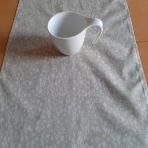 Könnte beinhalten: Eine weiße Keramik-Teetasse mit einem einzigartigen Griff liegt auf einem beigen Tischläufer. Der Läufer zeigt ein sich wiederholendes Muster aus weißen Hasen, Blumen und Eiern. Die Szene deutet auf ein Frühlings- oder Osterthema hin.