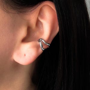 May include: A silver ear cuff with a flowing, organic design. The cuff wraps around the ear's cartilage, creating a modern and minimalist look. The jewelry is made of a shiny, reflective metal.