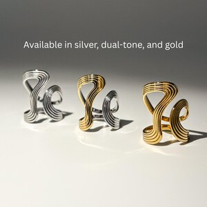 May include: Three abstract rings in silver, dual-tone, and gold. The rings have a unique, wavy design with parallel lines. The text "Available in silver, dual-tone, and gold" is displayed above the rings.