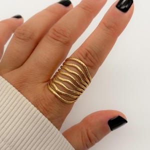 May include: A gold-colored ring with a textured, wavy design. The ring features multiple bands that wrap around the finger, creating a layered effect. The ring is worn on a finger with a black nail polish.