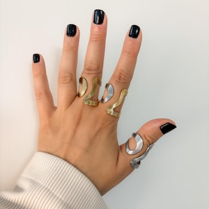 May include: A hand displaying several abstract wave-shaped rings. The rings are in gold and silver tones, with a minimalist design. The rings are worn on multiple fingers, showcasing the jewelry's unique style.