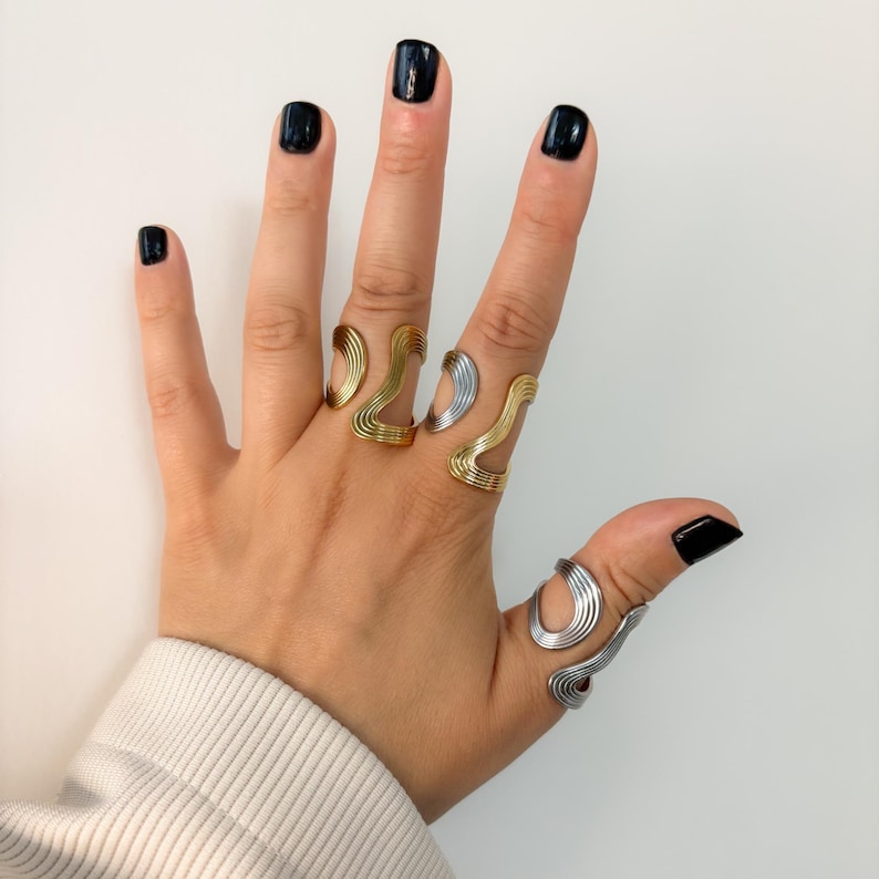 May include: A hand displaying several abstract wave-shaped rings. The rings are in gold and silver tones, with a minimalist design. The rings are worn on multiple fingers, showcasing the jewelry's unique style.