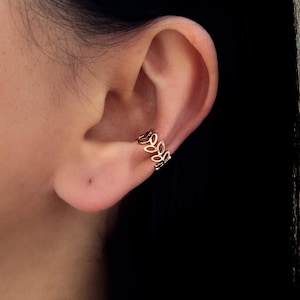 May include: A gold-colored ear cuff featuring a delicate leaf design. The cuff wraps around the ear's cartilage, creating a stylish and minimalist look. The openwork design allows light to pass through, enhancing its visual appeal.