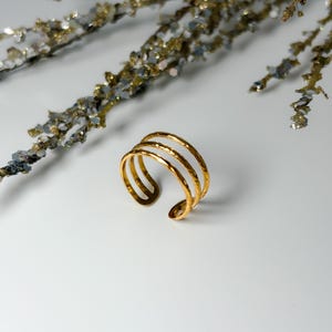 May include: A gold-colored cuff bracelet with a hammered texture. The open cuff design features three parallel bands. The jewelry is displayed on a white surface with decorative branches in the background.