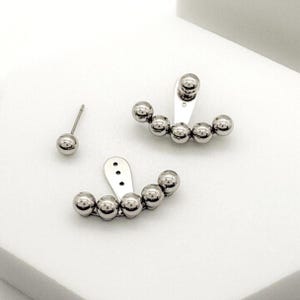 May include: Silver-toned earrings set. One earring is a simple stud, while the other two feature a curved design of five small spheres. The earrings are displayed on a white surface, showcasing their reflective quality.