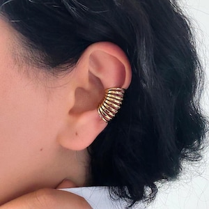 May include: A gold-colored ear cuff with a spiral design. The cuff wraps around the ear, creating a layered effect. The jewelry is worn on the ear. The background is blurred, focusing attention on the ear cuff.