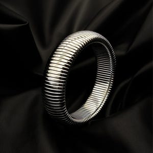 May include: A silver-coloured, ribbed bangle bracelet. The bracelet is circular and made of a series of parallel, horizontal lines. The jewellery is set against a black background, creating a contrast that highlights the bracelet's metallic sheen.