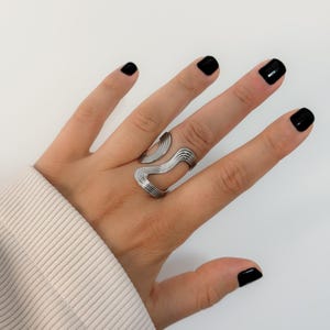 May include: A silver-colored, adjustable ring with a wavy design. The ring is worn on a hand with black nail polish and a beige sleeve.