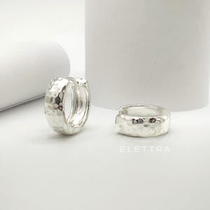 May include: Two silver-colored hoop earrings with a hammered texture. One earring is open, while the other is closed. The earrings are on a white surface with the word "ELETTRA" printed in the bottom right corner.