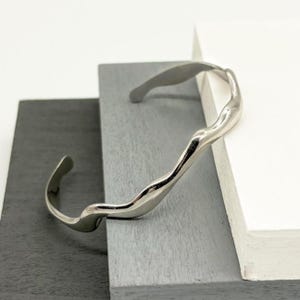May include: A silver-toned, open cuff bracelet with an organic, flowing design. The bracelet has a textured, wavy surface and a polished finish. The ends of the bracelet curve inward, creating a comfortable fit. The bracelet is displayed on a gray and white background.