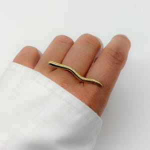 May include: A gold-colored ring with a wavy, elongated design. The ring is worn on a finger, and the background is a white shirt and a white background. The ring's design is smooth and minimalist.