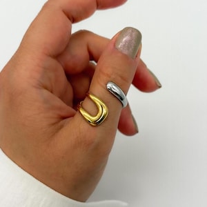 May include: A gold and silver ring set. The gold ring has a U-shaped design, while the silver ring wraps around the finger. The rings are made of shiny metal and are displayed on a hand.