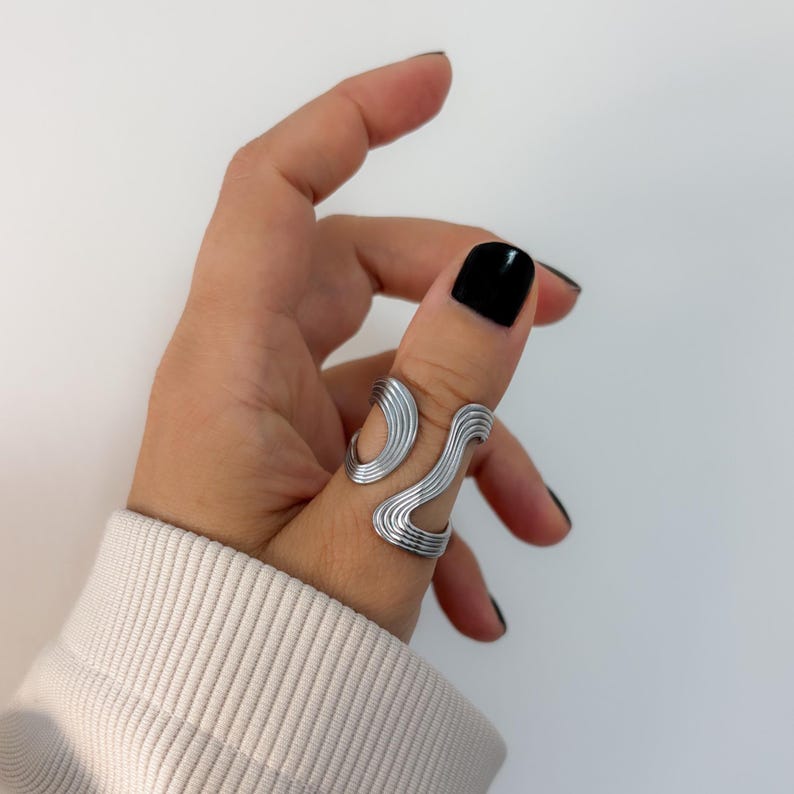 May include: A silver-colored ring with an abstract, wavy design. The ring wraps around the finger with a series of parallel lines, creating a modern and artistic look. The ring is worn on a finger with black nail polish.
