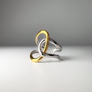 May include: A modern ring with a unique design. The ring features a gold-colored loop intertwined with silver-colored loops. The ring is made of metal and has a polished finish. The ring is a piece of jewelry.