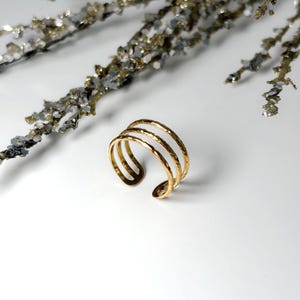 May include: A gold-colored cuff bracelet with three textured bands. The open-ended design allows for easy wear. The bracelet is set against a white background with decorative silver and gold-toned branches.