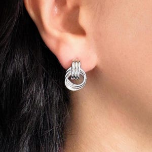 May include: A silver earring with a circular design is shown. The earring has multiple interlocking rings, creating a textured and modern look. The earring is attached to an ear, and the overall aesthetic is minimalist and elegant.
