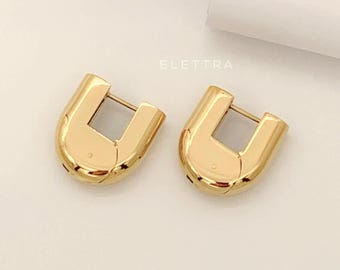 May include: Two gold-colored, U-shaped hoop earrings. The earrings have a polished finish and a rectangular opening in the center. The word "ELETTRA" is printed in a light color.