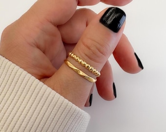 May include: Two gold rings are worn on a finger. One ring is a twisted band, and the other is a beaded band. The rings are a bright gold color. The background is a light color.